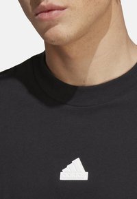 T-shirt nera in cotone con scollatura rotonda a costine e logo Adidas bianco testurizzato sulla parte sinistra del petto.