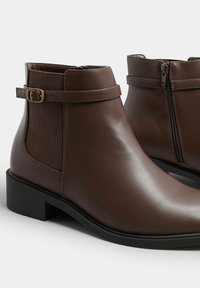 Botas de piel marrón hasta el tobillo con un panel lateral texturizado, correa ajustable y cremallera lateral. Punta cuadrada y tacón bajo en bloque.