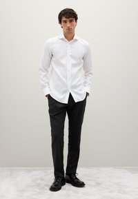 Camicia bianca a maniche lunghe con colletto classico, abbinata a pantaloni scuri a righe sottili. Scarpe di pelle nere completano l'outfit.