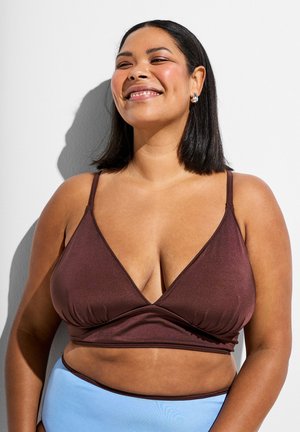 Smilende kvinde iført en brun bralette og lyseblå højttaljede underdele mod en hvid baggrund med skygger.