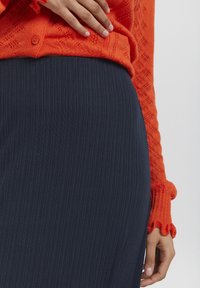 Oranssi neulecardigan, jossa on teksturoitu kuvio, nappidetaili, yhdistetty tummansiniseen ribbihameeseen. Materiaali näyttää pehmeältä ja joustavalta.