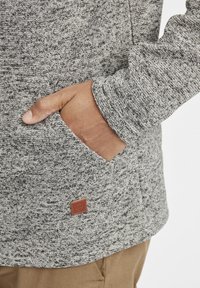 Gray textured sweatshirt with a pocket on the side. Features a brown leather brand tag at the seam. Soft, casual fabric.