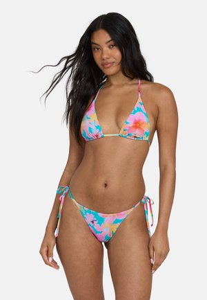 Vrouw met lang donker haar die een kleurrijke bloemige tie-dye bikini draagt met een driehoekstop en zijsstrik broekje, staand tegen een effen achtergrond.