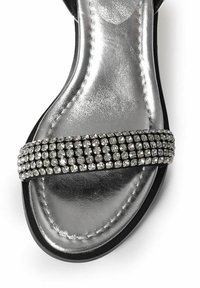 Sandalia negra con una amplia tira adornada con pequeñas y brillantes piedras de strass. La plantilla es de color plateado metálico con una textura suave.