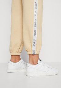 Cream-colored joggers with elastic cuffs feature a side stripe with "LEVI'S 1873" in white. Paired with white Nike sneakers.