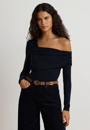 Femme aux longs cheveux bouclés portant un haut noir à une épaule avec manches longues et un pantalon noir avec une ceinture marron, debout avec une main dans la poche.