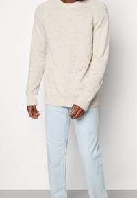 Ljusbeige stickad tröja med texturerad yta, ribbade ärmslut och prickiga accenter. Matchad med ljusblå raka jeans.