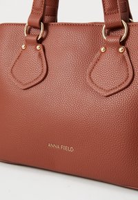 Sac à main texturé marron avec deux poignées, accents dorés, et un logo en relief sur le devant. Présente un design élégant et structuré.