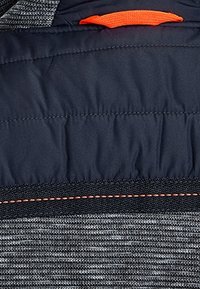 Tmavá polstrovaná část z textilie s horizontálním stehováním, nad šedou texturovanou pleteninou, obsahující oranžový štítek a černý popruh s oranžovým šitím.