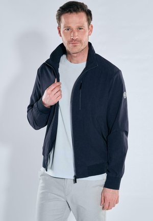 Man wearing navy zip-up jacket over white shirt and light-colored pants, adjusting jacket collar, plain light background.