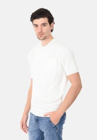 Maglietta bianca a maniche corte in cotone, con una singola tasca sul petto, scollatura rotonda e polsini a coste, abbinata a jeans blu.