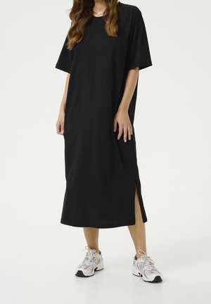 Frau trägt ein lockeres schwarzes Midikleid mit kurzen Ärmeln und seitlichem Schlitz, kombiniert mit weißen Sportsneakern, steht vor weißem Hintergrund.