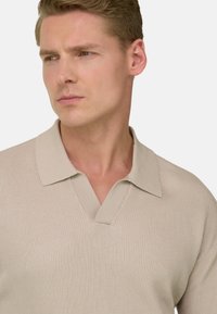 Jeune homme aux cheveux châtain clair portant une chemise à col en maille côtelée beige, regardant vers sa gauche sur un fond uni.