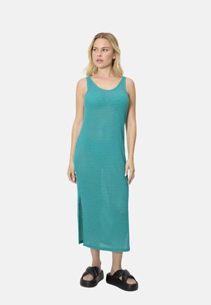 Mujer de pie con un vestido midi de punto azul verdoso sin mangas, con aberturas laterales y sandalias negras de plataforma, sobre un fondo claro y liso.