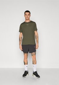 Olivengrønn T-skjorte med korte ermer og hvit merkevare, kombinert med svarte treningsshorts. Svarte joggesko og hvite sokker fullfører antrekket.
