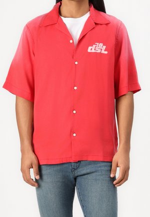 Homme portant une chemise à manches courtes rouge vif avec un logo blanc sur la poitrine, associée à un jean bleu clair.