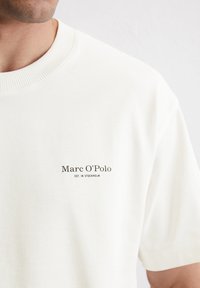 Short-sleeve, cream-colored cotton t-shirt featuring a ribbed neckline and a printed logo with "Marc O'Polo" and "EST. IN STOCKHOLM."