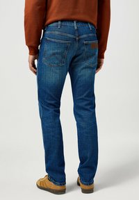 Vaqueros de mezclilla azul con un corte recto, bolsillos traseros clásicos con un detalle de costura estampada y una etiqueta de cuero marrón en la cintura.