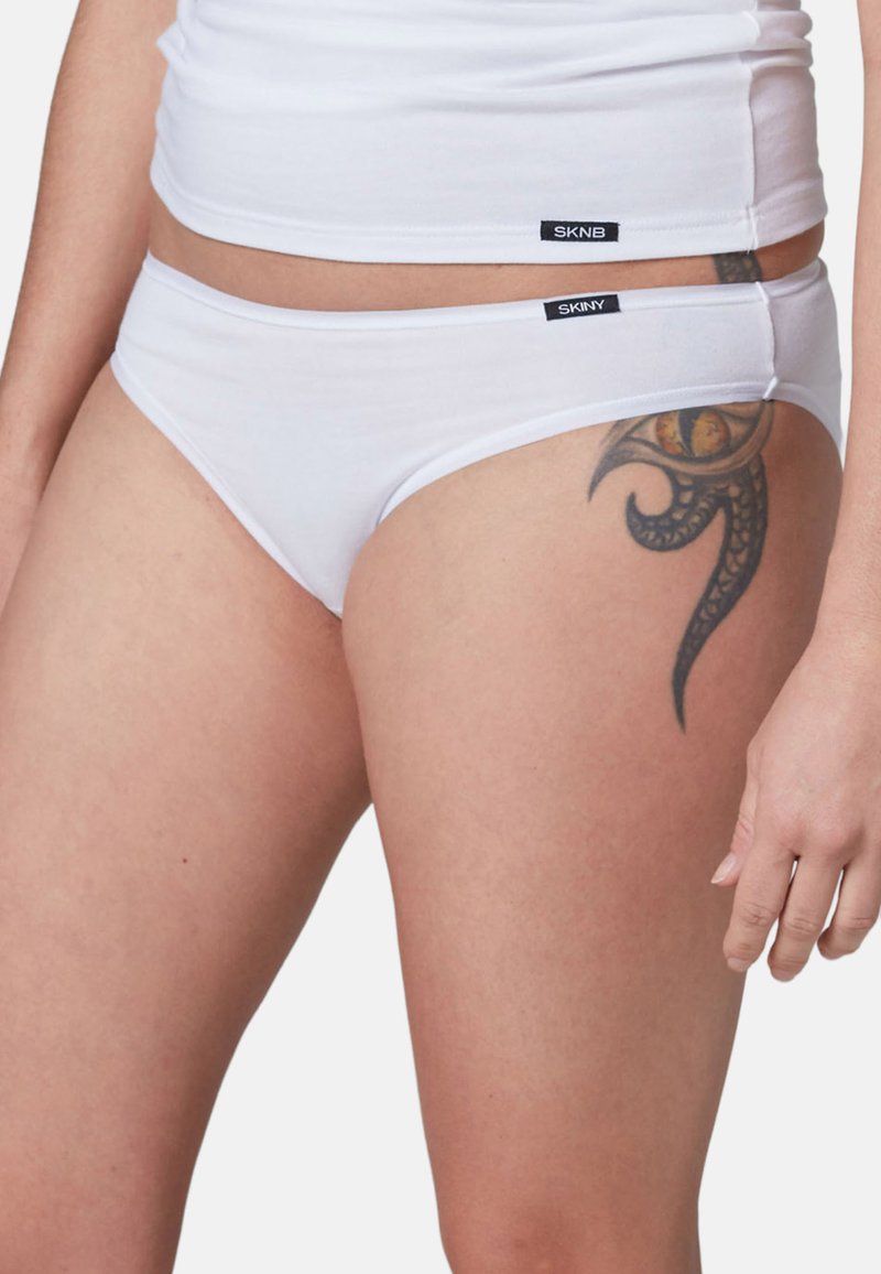 Nahaufnahme einer Person, die weiße Unterwäsche und ein Tanktop trägt, mit einem schwarzen und orangefarbenen Tribal-Tattoo am oberen Oberschenkel.