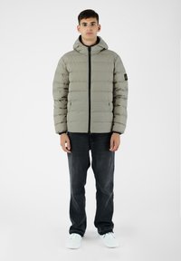 Giacca a vento grigia chiara con chiusura a zip e cappuccio, caratterizzata da un design aderente. Abbinata a jeans scuri e sneakers chiare.