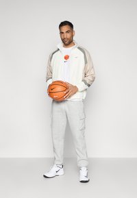 Λευκό αθλητικό μπουφάν με γκρίζα μανίκια από δίχτυ, γκρίζο φούτερ με cargo τσέπες, λευκά αθλητικά παπούτσια με μαύρες λεπτομέρειες, κρατώντας ένα πορτοκαλί μπάλα μπάσκετ.