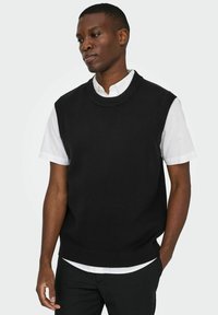 Schwarzer gestrickter ärmelloser Pullover mit rundem Halsausschnitt, strukturiertem Stoff und geripptem Saum. Getragen über einer weißen Bluse mit Kragen.