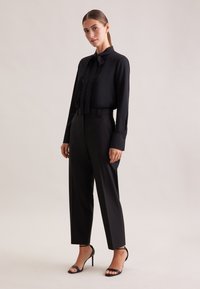 Blusa nera con fiocco al collo e dettagli in pizzo, abbinata a pantaloni neri a vita alta e sandali neri con tacco, su sfondo neutro.