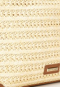 Tkaná slaměná taška světlé béžové barvy s texturovaným povrchem. Obsahuje hnědou koženou nášivku s názvem značky a charakteristickými tkanými vzory.