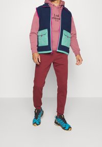 Marineblaues und teal-farbenes Vest mit zwei vorderen Taschen, über einem rosa Hoodie getragen, kombiniert mit burgunderroten Hosen und blauen Sneakers mit schwarzen Akzenten.