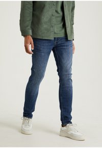 Vaqueros de mezclilla azul con un corte slim, que presentan un lavado medio y un leve desgastado. Combinados con zapatillas blancas y una chaqueta de camisa de pana verde.