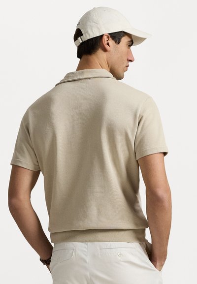 Polo shirt beige à manches courtes avec une texture côtelée et un col rabattu, associé à un pantalon blanc et une casquette de couleur claire.