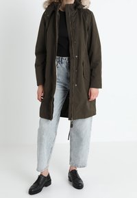 Parka verde olivo con capucha forrada de piel, que cuenta con un cierre delantero, botones a presión y dos bolsillos laterales. Lleva puesta una blusa negra y jeans claros.