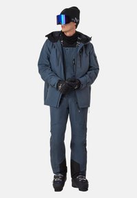 Dunkelblaue isolierte Skijacke mit passenden Hosen, ausgestattet mit mehreren Taschen und Reißverschlüssen. Schwarze Handschuhe und Beanie, große verspiegelte Skibrille.