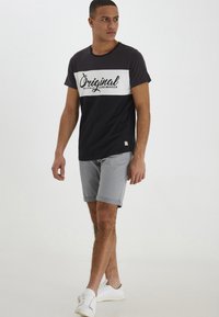 T-shirt noir et gris avec une bande horizontale blanche portant le texte "Original" en écriture manuscrite. Associé à un short gris clair et des baskets blanches.