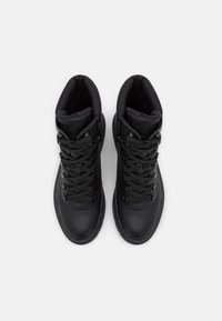 Une paire de bottes d'hiver en cuir noir avec des cols rembourrés, des œillets en métal et de grosses lacets noirs, vues de dessus sur un fond blanc.