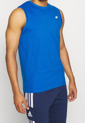 Mężczyzna ubrany w niebieską koszulkę bez rękawów oraz granatowe spodnie Adidas z trzema białymi paskami po bokach, stoi na neutralnym tle.