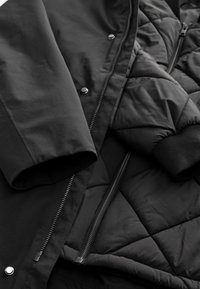 Gros plan sur une veste matelassée noire portée sous un manteau noir lisse, montrant les manches, les fermetures éclair et les boutons-pression.