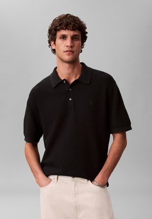 Ung mand med krøllet hår iført en sort kortærmet strikket poloshirt og lyse beige bukser, hænderne i lommerne, neutral grå baggrund.
