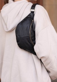 Bolso de sling de cuero negro con un bolsillo con cremallera, forma curva y correa ajustable, llevado sobre una sudadera de color claro.