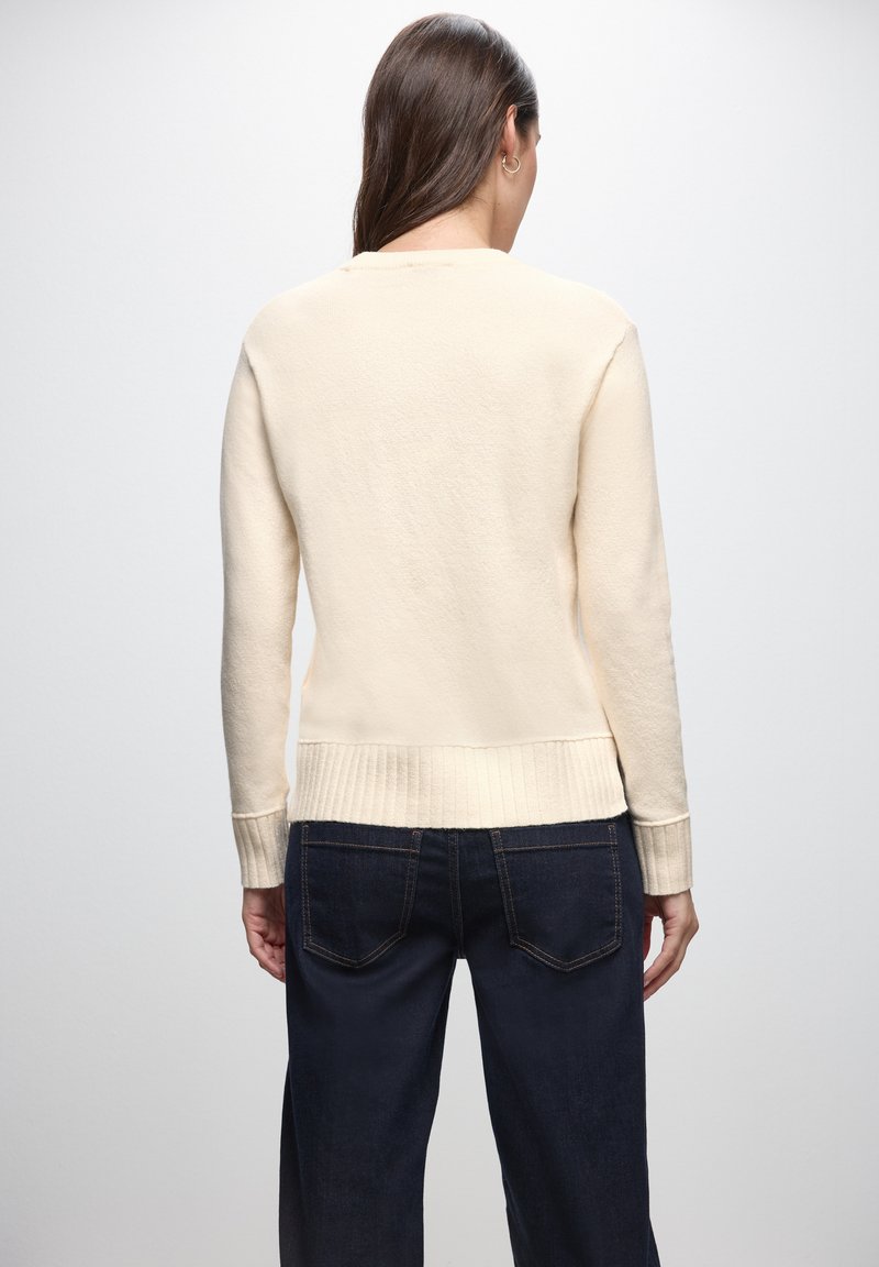 Cremfarbener Strickpullover mit geripptem Saum und Bündchen, im entspannten Schnitt und mit glatter Textur. Kombiniert mit dunkelblauen Denim-Jeans.
