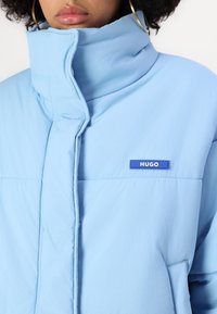Žena v světle modré vatované bundě s vysokým límcem a malým tmavě modrým logem "HUGO" na hrudi, viditelné zlaté kruhové náušnice.