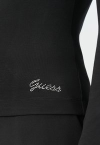 Czarny materiał z małym logo "Guess" wyłożonym przezroczystymi cyrkoniami blisko dolnego brzegu długiego rękawa odzieży.