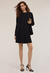 Abito nero senza maniche con una silhouette svasata, abbinato a un blazer nero sartoriale e sandali neri con cinturini. Tiene una borsa a tracolla con texture.