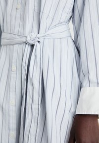 Gaiši zils krekla kleitas modelis ar vertikālām tumši zilo svītrām, garām piedurknēm, jostiņas viduklī, pogu aizdari priekšpusē un baltām aprocēm. Gluda auduma tekstūra.