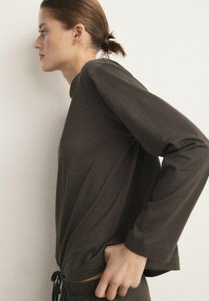 Woman with tied-back hair wearing a dark long-sleeve top and matching pants, standing in profile against a light wall.