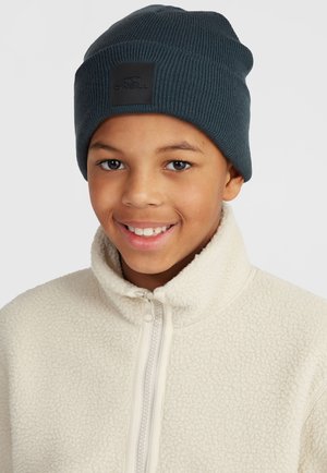 Donkerblauwe ribgebreide beanie met een vierkant zwart logo patch. Draagt samen met een crèmekleurige fleece jack met een rits.