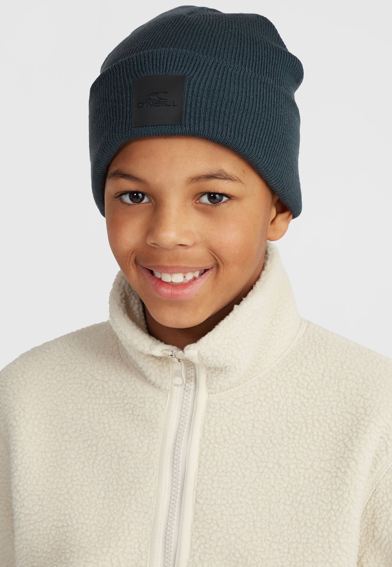 Berretto a costine blu scuro con una patch quadrata nera. Indossato con una giacca di pile color crema con zip.