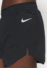 Lähikuva mustista Nike urheilushorteista, joissa on vetoketjullinen tasku ja valkoinen Nike-logo henkilön reidellä.