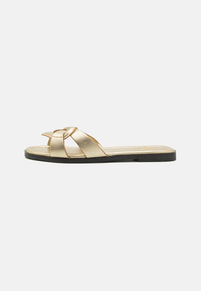 Sandal de couro dourado com tiras cruzadas, sola plana preta e detalhe de costura subtil. Textura suave com acabamento metálico.
