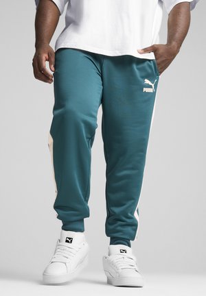 Turkusowe spodnie sportowe wykonane z gładkiego materiału z białymi paskami po bokach, w połączeniu z białymi sneakersami i luźną białą koszulką. Widoczne logo PUMA.